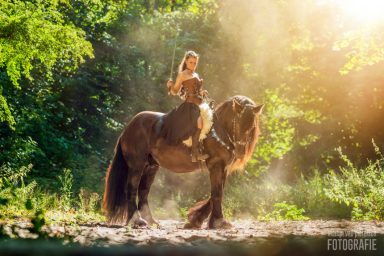 Fotoshoot met paard Een vrouw in een lichtbruin kostuum zit op een paard in een bosrijke omgeving.