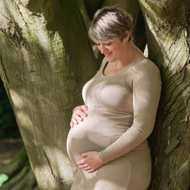 Zwangerschapsfotoshoot Arboretum Heempark Delft Zwangere vrouw in een beige jurk leunt tegen een boom en kijkt naar haar buik.