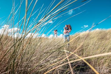 Fotoshoot in de duinen Twee mensen lopen door hoge grassprieten onder een heldere blauwe lucht.