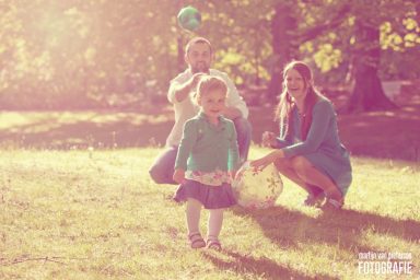 Gezin fotoshoot op landgoed Clingendael Kind met een mandje in een zonnige tuin, geflankeerd door een vrouw en een man.