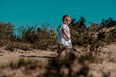 Fotoshoot in het Westduinpark Een klein meisje in een witte jurk wandelt langs een zandpad met groene planten op de achtergrond.