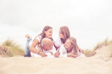 Fotoshoot met het hele gezin in het Westland Een gezin met vier personen ligt lachend op een zandige bodem bij struiken.