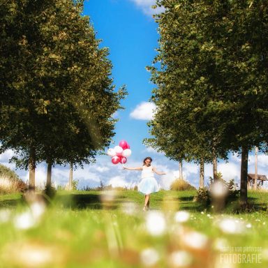 Fotoshoot in je eentje Een meisje in een witte jurk rent met ballonnen door een zonnig grasland.