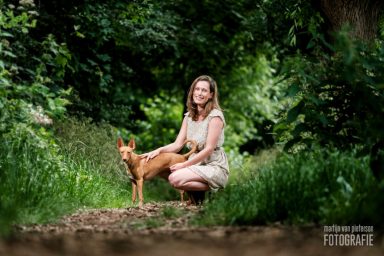 Fotoshoot Prinsenbos in Naaldwijk Een vrouw in een lichte jurk zit op een pad, naast een bruin hondje, omringd door groen.