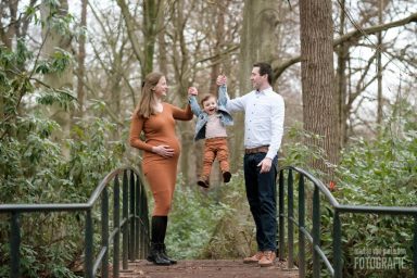 Zwangerschapsfotoshoot in de winter Een zwangere vrouw en een man houden een kind vast boven een brug in een bosrijke omgeving.